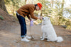 Man in a brown jacket and red cap interacting with a white dog on a forest path.